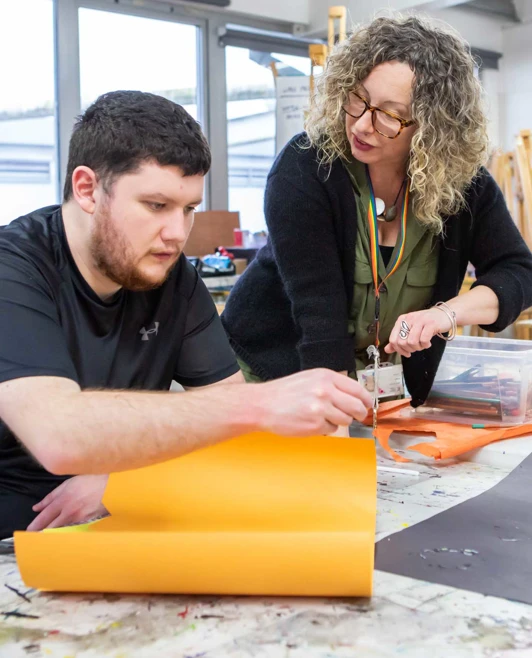 Lecturer assisting a student with an art project, pointing at a sheet of orange paper, while standing in a classroom filled with easels and supplies. Lecturer assisting a student with an art project, pointing at a sheet of orange paper, while standing in a classroom filled with easels and supplies.
